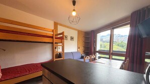 A compact room with a bunk bed, a small kitchenette, a dining area, and a large window offering a scenic view.