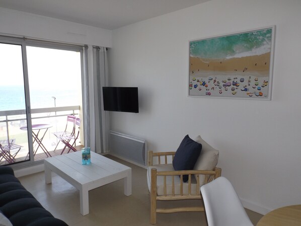 Un salon avec un canapé blanc, une chaise en bois, une table basse blanche et un téléviseur fixé au mur.