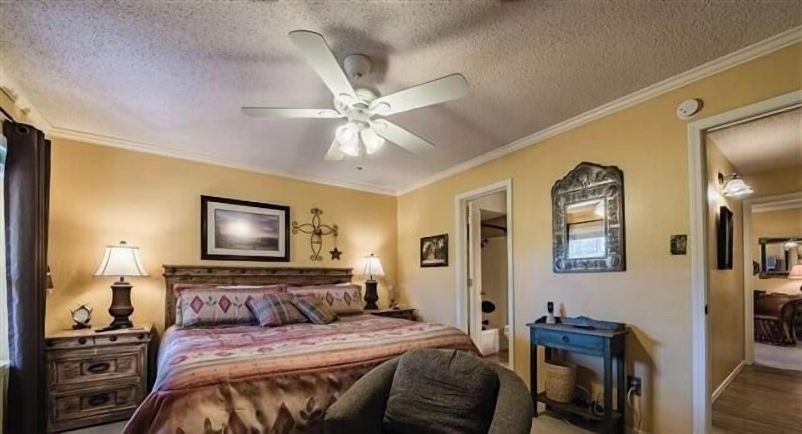 A bedroom with a bed, nightstands, a ceiling fan, a mirror, and a door leading to another room.