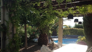 Un patio couvert avec une table et des chaises, entouré de verdure et donnant sur une piscine.