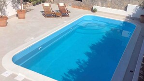 Una piscina rectangular con borde blanco, rodeada por una terraza con baldosas. Hay dos sillones de madera y varias plantas en macetas alrededor de la piscina.