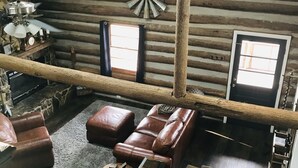 Interior de una cabaña de troncos con escalera de madera, asientos de cuero, chimenea y decoración con cabeza de venado.