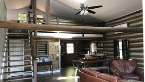 Interior rústico de una cabaña con escalera de madera, comedor, sofá de cuero y ventilador de techo.