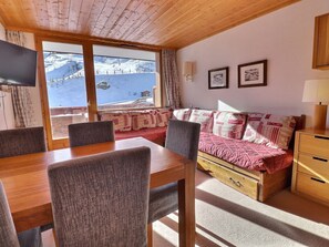 Une pièce chaleureuse avec une table et des chaises en bois, un canapé agrémenté de coussins à motifs, un meuble en bois et une fenêtre donnant sur un paysage de montagne enneigée.