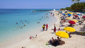 Una playa con aguas turquesas cristalinas, gente nadando y tomando el sol, y sombrillas coloridas.
