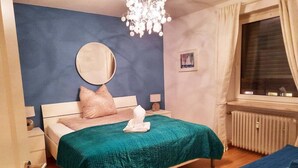 A bedroom with a bed, a bedside table, a nightstand, a mirror, a window with curtains, and a radiator.