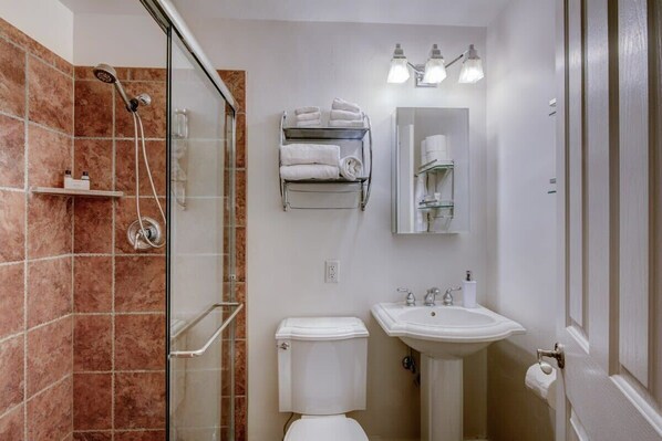 Een badkamer met een douchecabine van glas, een wit toilet, een wastafel met een spiegel erboven en planken met handdoeken.