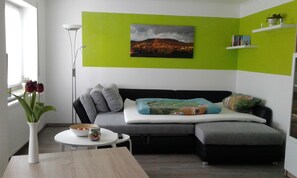 A modern living room with a grey sofa, a green accent wall, a small table with a vase of flowers, and a painting of a mountain scene.