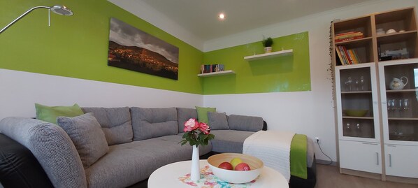 A living room with a grey sofa, a white table with a bowl of fruit, a green wall, a bookshelf, and a cabinet with glass doors.