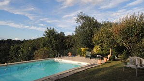 A rectangular swimming pool with a metal ladder, surrounded by a grassy area and a metal fence. There are potted plants and a wooden bench near the pool. - In a listed village, half-timbered house overlooking a wooded valley (Montjoi)
