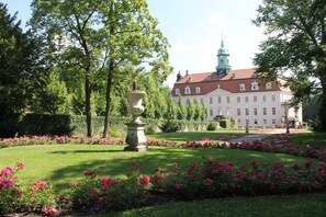 Ein großes Gebäude mit einem Turm, umgeben von einem gepflegten Garten mit rosa Blumen und einer zentralen Fontäne.