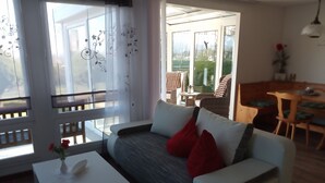 Een woonkamer met een witte bank, rode kussens, een witte salontafel en een vaas met rode bloemen.