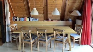 Un coin repas en bois, avec une longue table et six chaises, deux lampes suspendues et une bibliothèque remplie de livres.