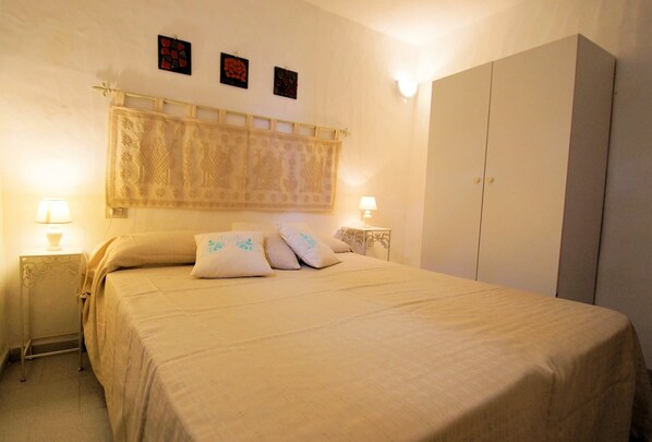 Una cama bien hecha con una colcha color beige y almohadas, dos lámparas de noche, un armario grande y un tapiz con motivos en la pared.