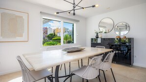 Un espace de restauration moderne avec une grande table, des chaises blanches, un lustre et une vue sur les bâtiments à travers de grandes fenêtres.