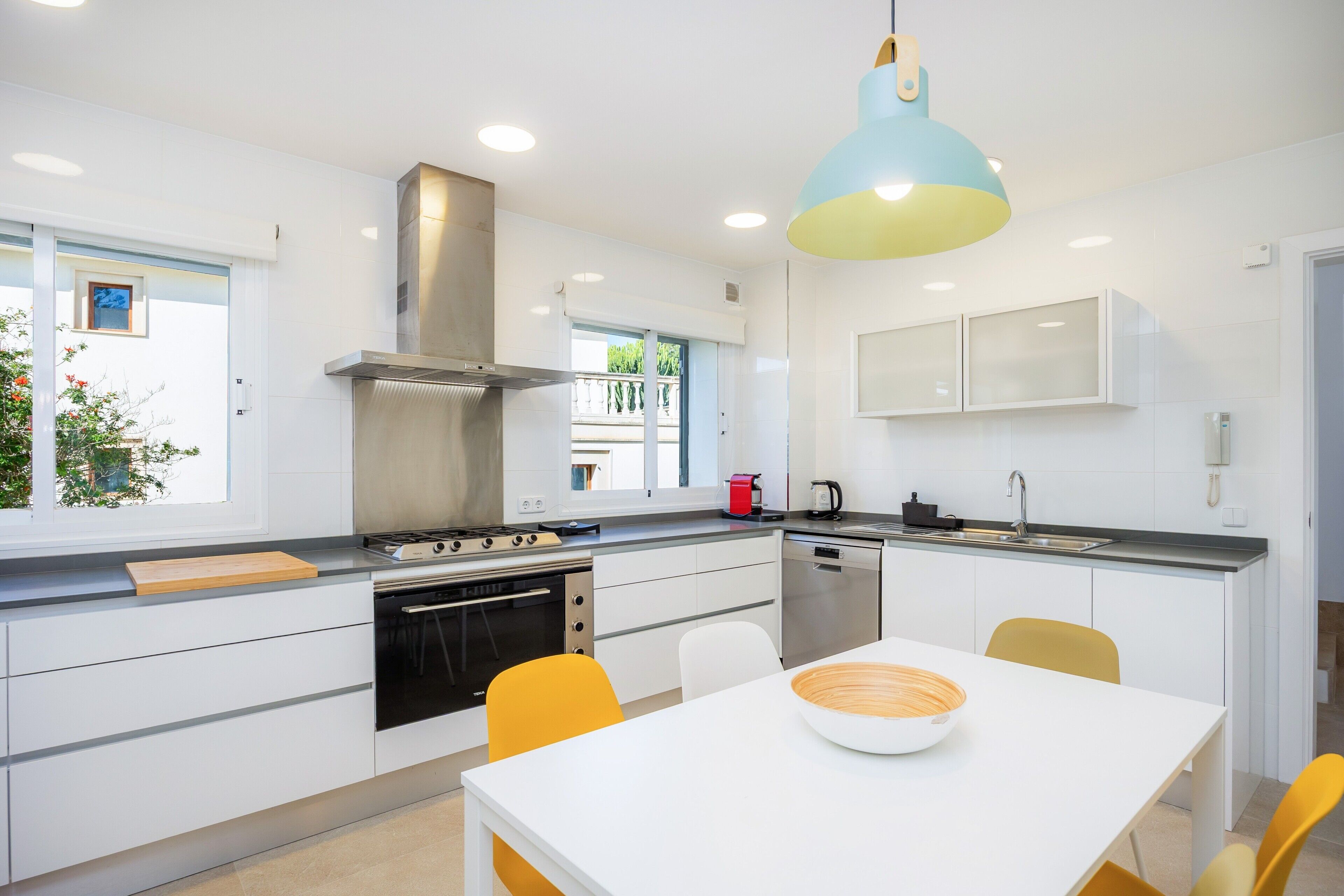 Una cocina moderna con muebles blancos, extractor de acero inoxidable, horno empotrado y comedor con mesa blanca y sillas amarillas.