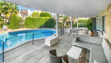 A covered outdoor swimming pool area with a curved pool, seating, and a view of the surrounding buildings and greenery.