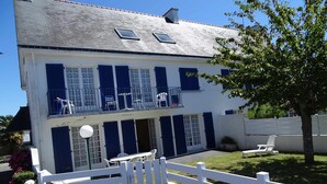 Un bâtiment à deux étages, doté d’une façade blanche et de volets bleus, avec un balcon aménagé en espace de détente extérieur et une entrée fermée par un portail.