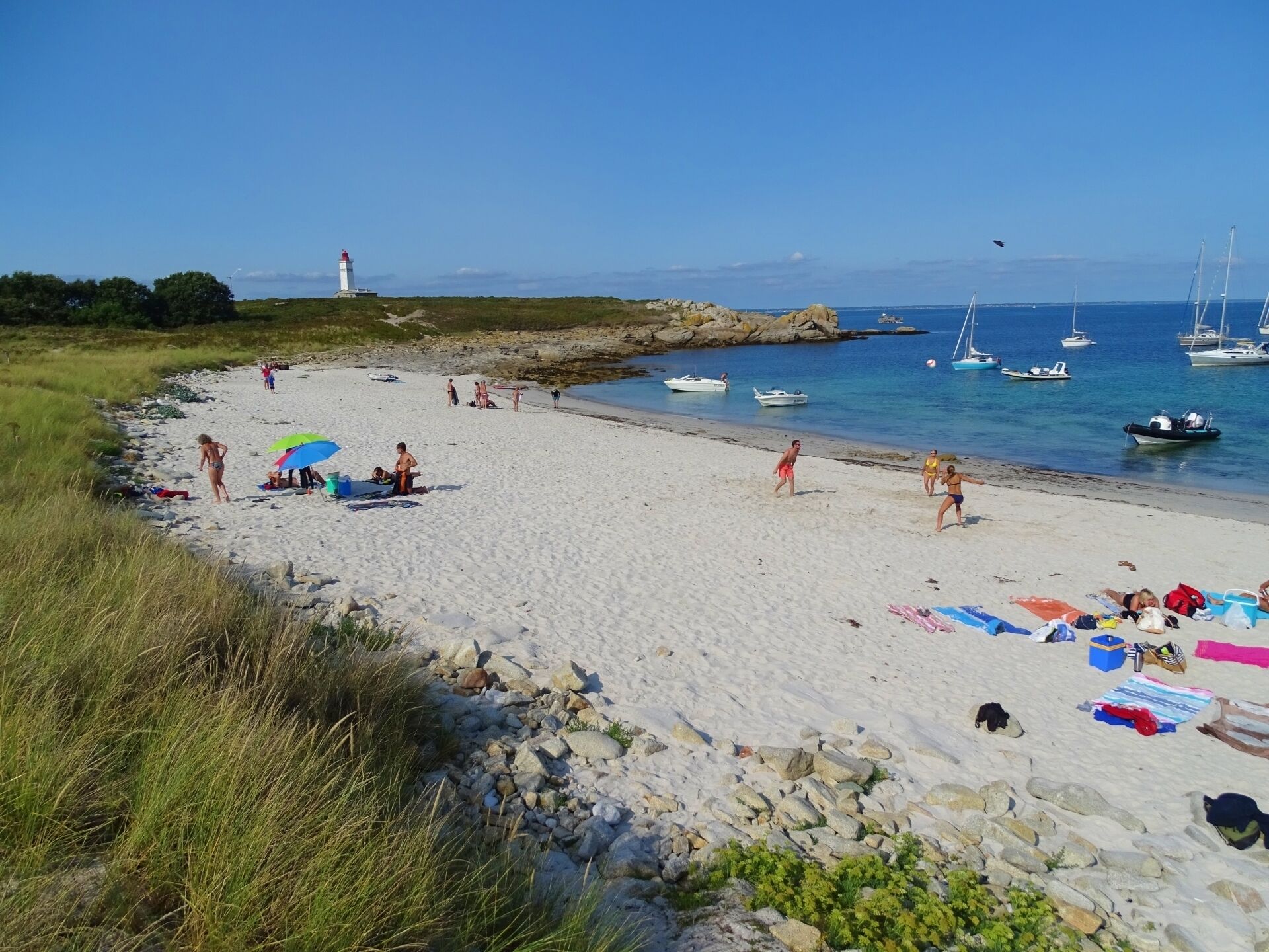 A beach with people sunbathing and swimming, boats in the water, and a lighthouse in the distance.