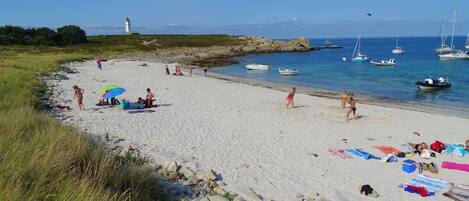 A beach with people sunbathing and swimming, boats in the water, and a lighthouse in the distance.