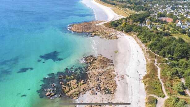 Vue aérienne d’une zone côtière avec une plage de sable, des affleurements rocheux et une zone résidentielle avec des maisons.
