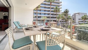 Un balcón con mesa lista para comer, rodeado de sillones de descanso y vistas a una ciudad con palmeras y un edificio de varios pisos.