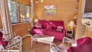 Una habitación acogedora con detalles de madera, un sofá rosa, sillas de mimbre, una mesita y un televisor sobre un mueble.