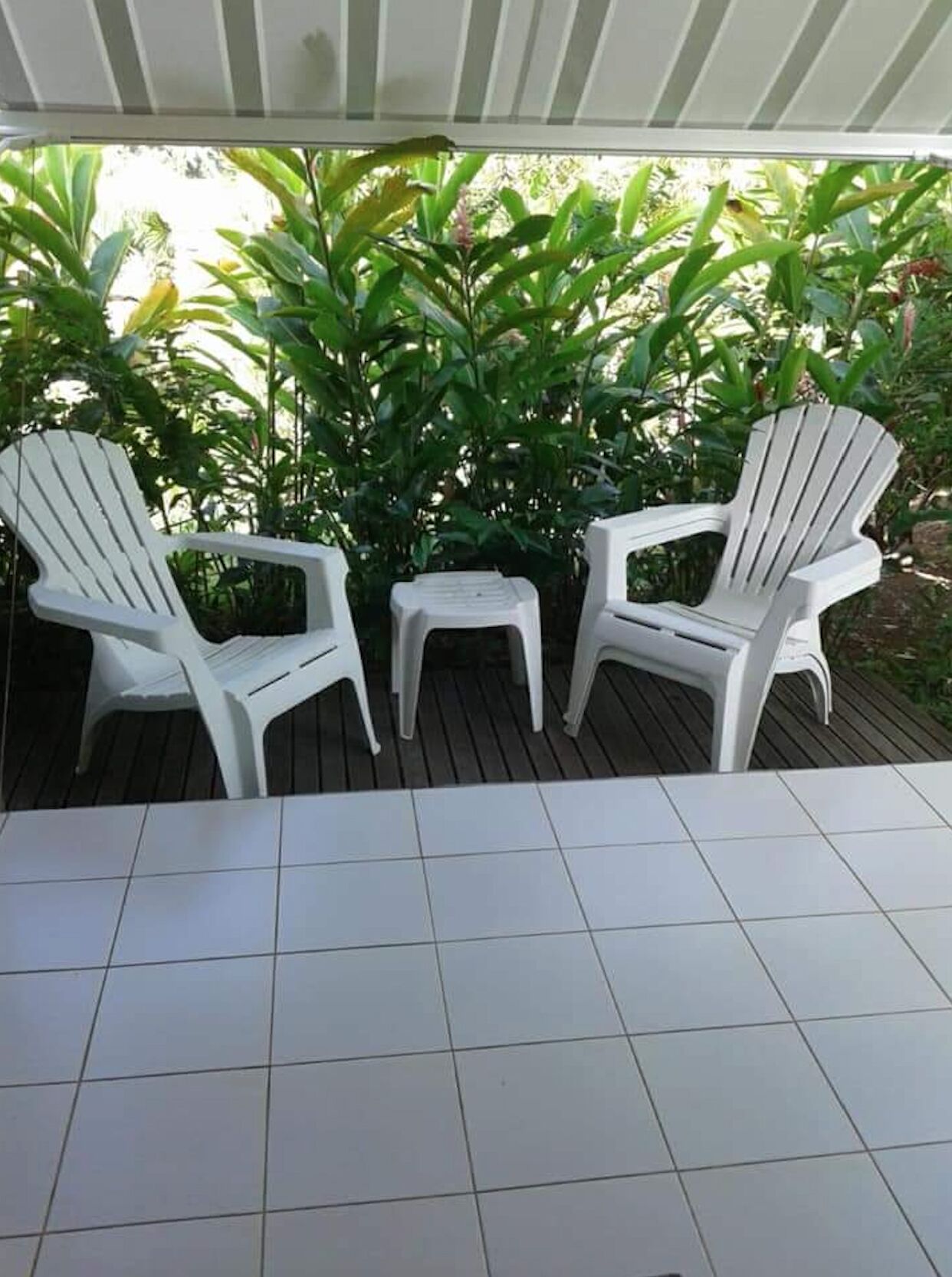 Three white plastic chairs and a small table on a tiled patio area with a wooden deck, surrounded by lush green foliage.