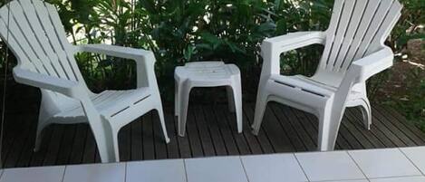 Three white plastic chairs and a small table on a tiled patio area with a wooden deck, surrounded by lush green foliage.