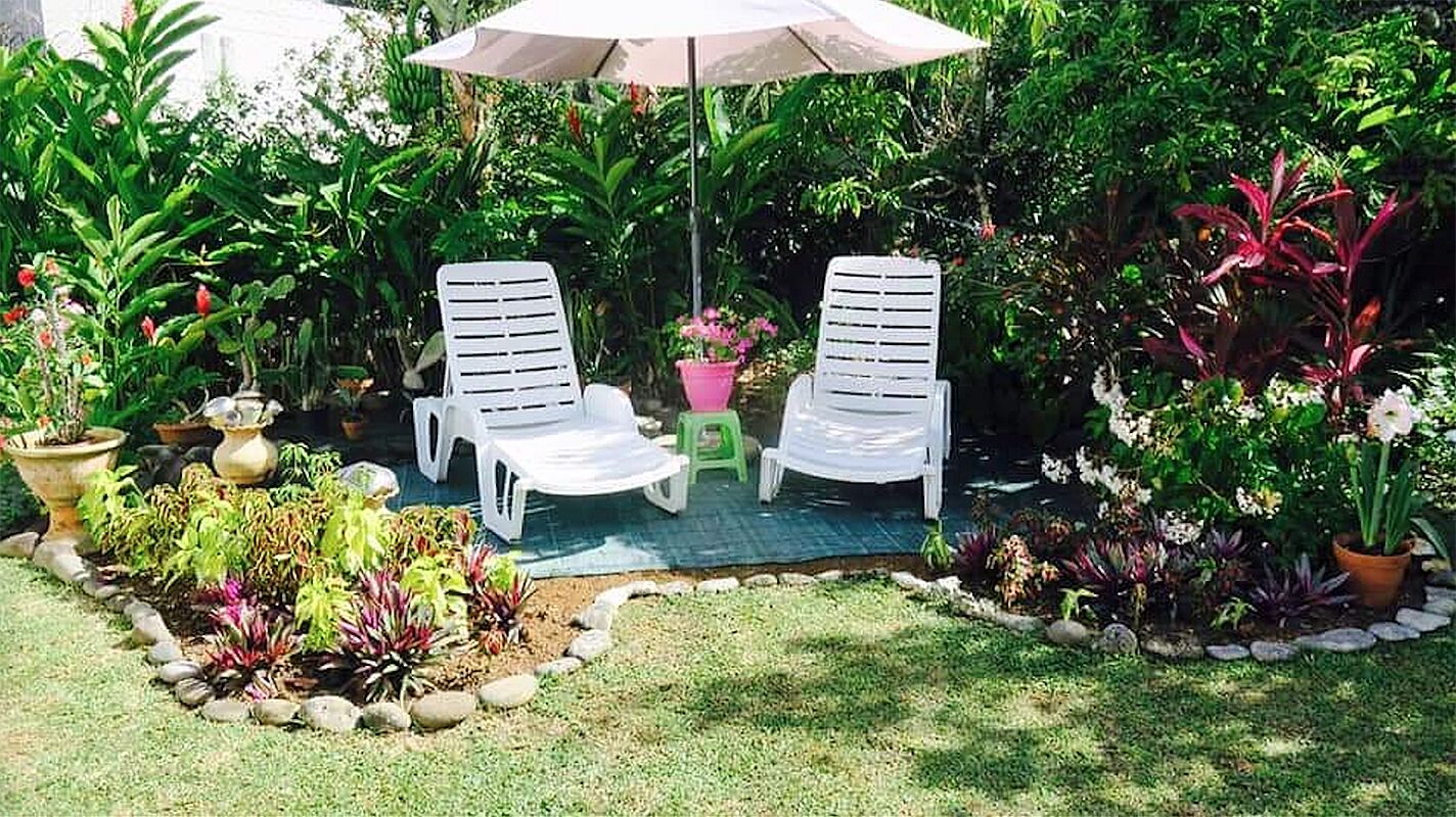 Zwei weiße Lounge-Stühle unter einem großen Sonnenschirm in einem Garten mit verschiedenen Kübelpflanzen und einem kleinen Hocker.