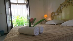 Een netjes opgemaakt bed met opgerolde witte handdoeken, een potplant met een rode bloem en een lamp op een nachtkastje.