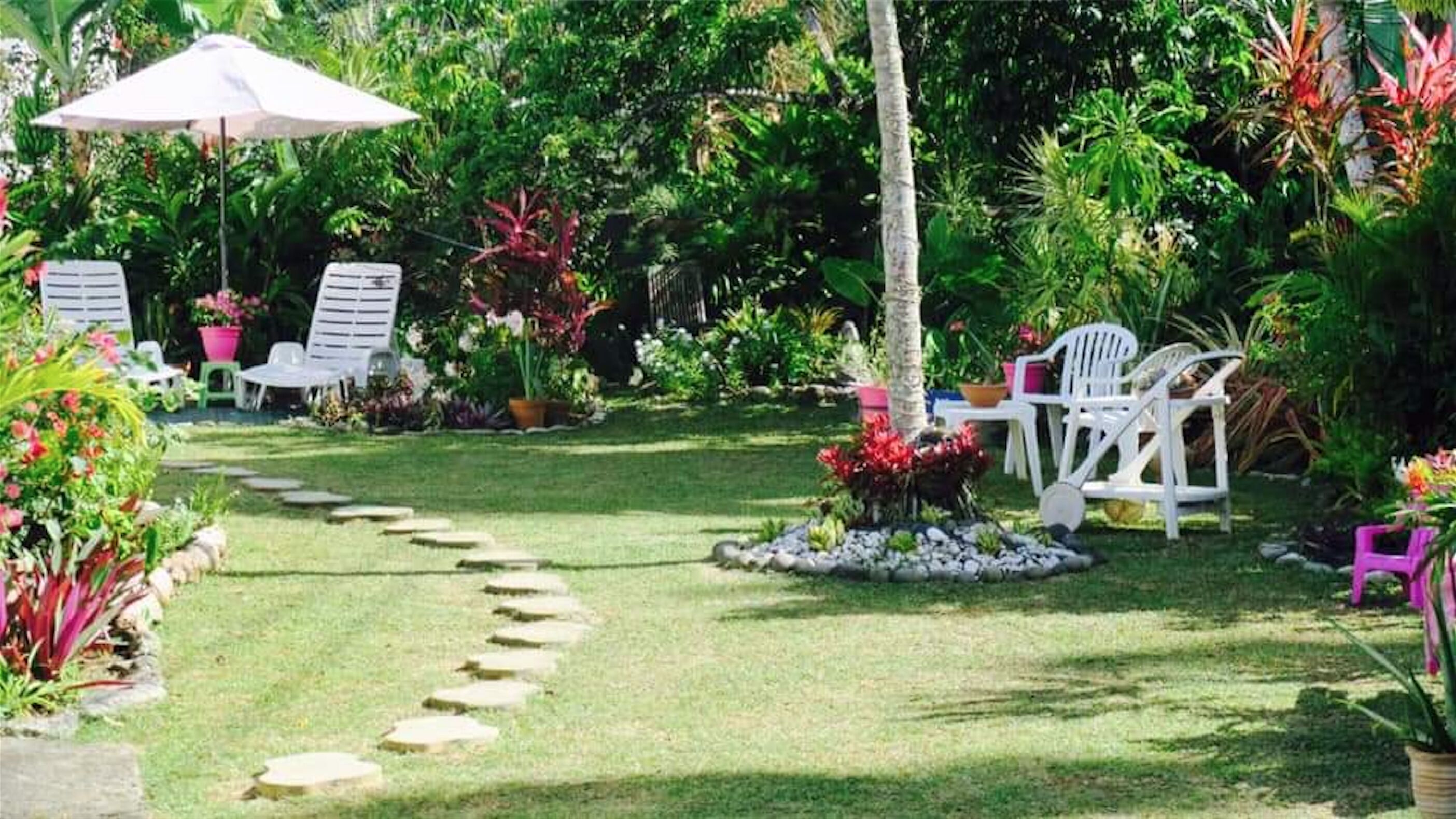 A garden with a stone path, white chairs, and a table under a large umbrella.