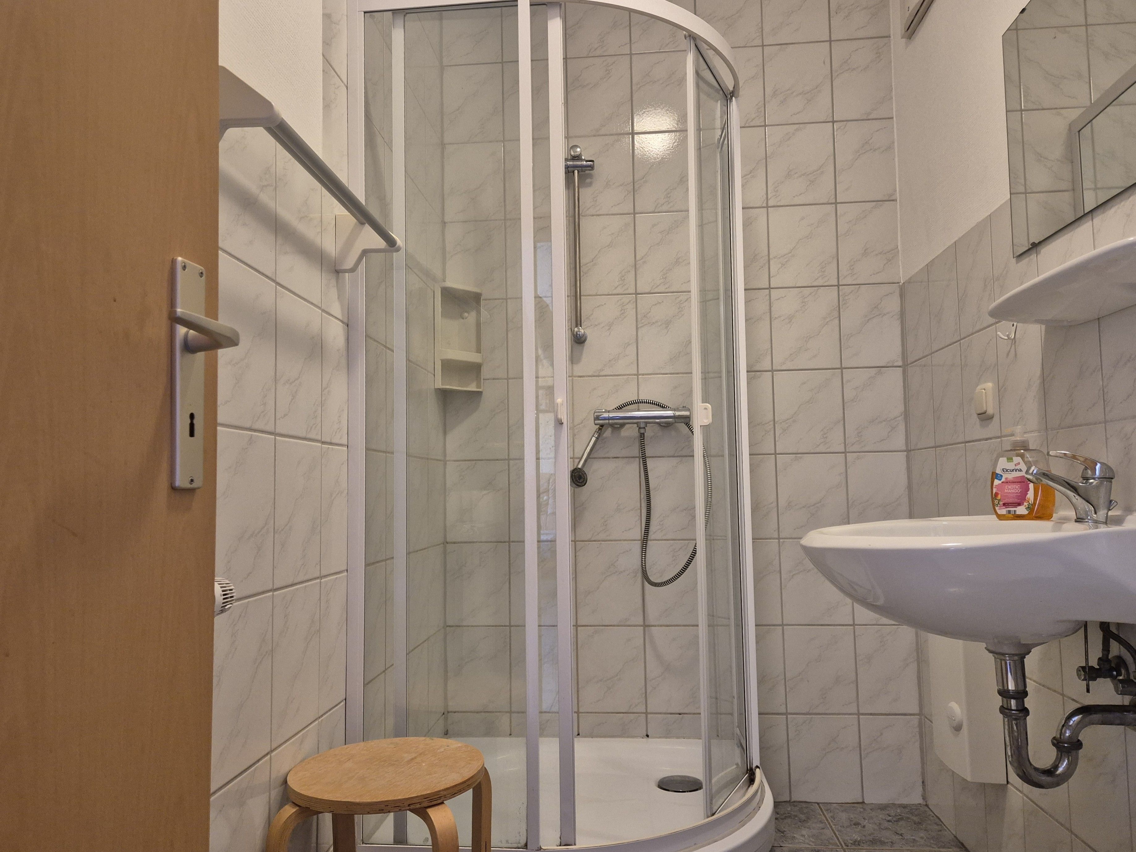 Une salle de bain avec une douche à parois de verre, un lavabo doté d’un distributeur de savon et un tabouret en bois.