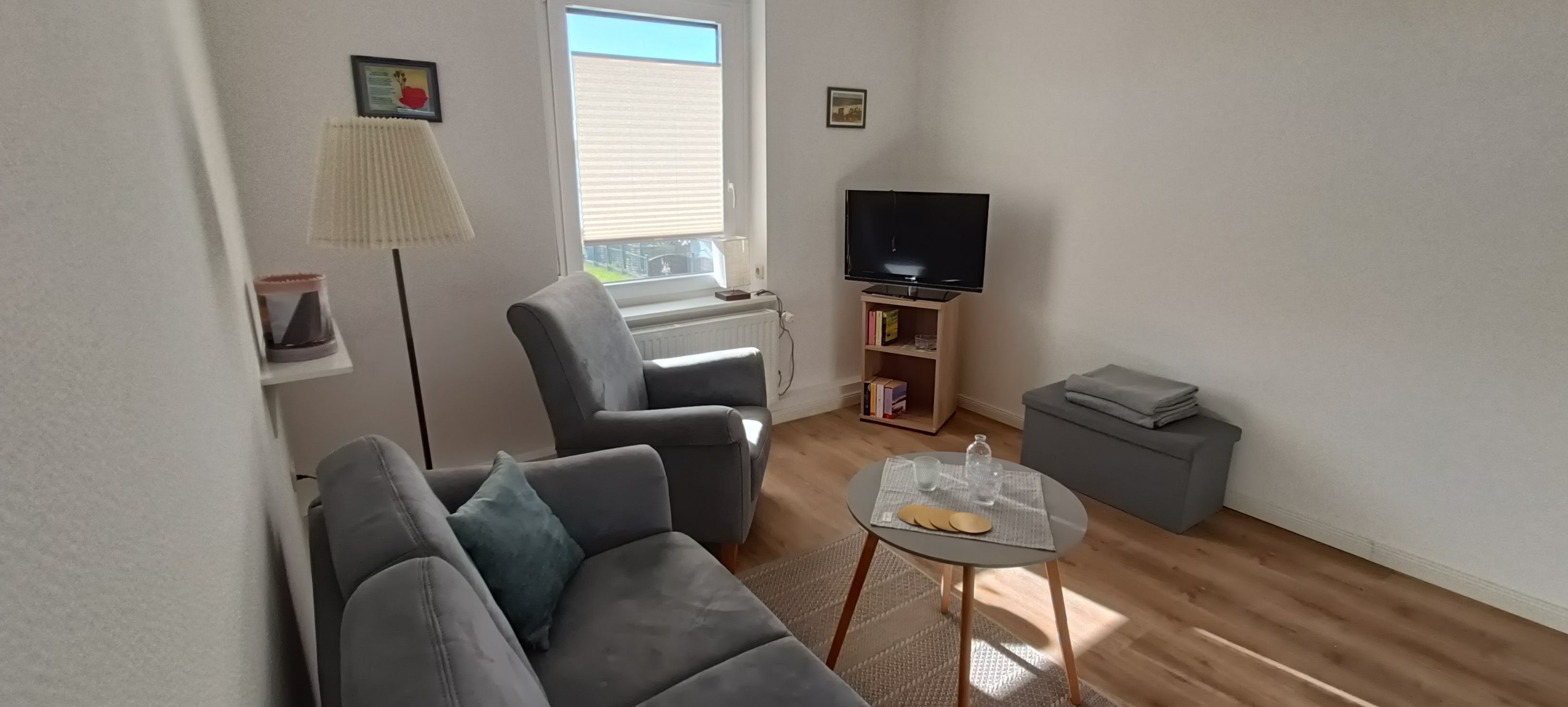 Un salon avec un canapé gris, une petite table ronde avec un plateau en verre, un lampadaire, un téléviseur posé sur un meuble et une bibliothèque.