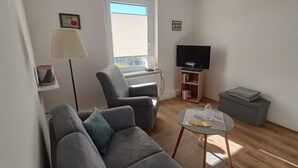 A living room with a grey sofa, a small round table with a glass top, a floor lamp, a television on a stand, and a bookshelf. - Apartment on the upper floor - apartment cicilie (Maasholm)