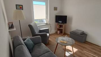 Un salon avec un canapé gris, une petite table ronde avec un plateau en verre, un lampadaire, un téléviseur posé sur un meuble et une bibliothèque.