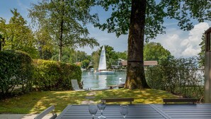 Une table avec trois verres à vin et un banc, donnant sur un lac où l’on aperçoit un voilier et une maison au loin.