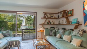 Un salon avec un canapé vert, un pouf à rayures et une table basse. On y trouve des étagères ornées d’objets décoratifs et une vue sur un voilier à l’extérieur.