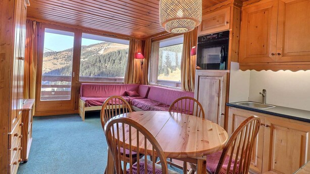 A wooden cabin interior with a dining table, chairs, a sofa, and a kitchen area.