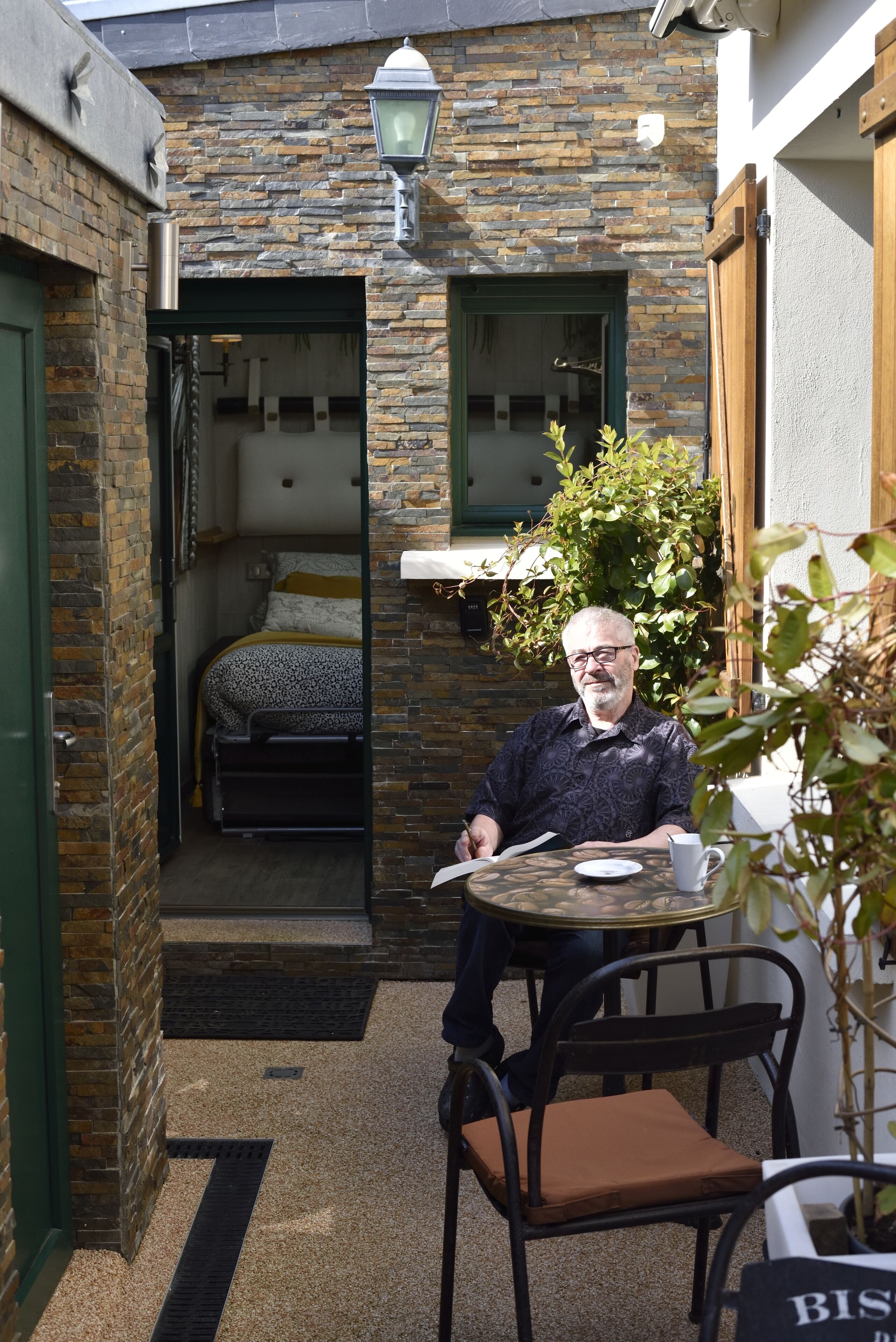 A man sitting at a table in a brick-walled outdoor area with a bed visible through an open doorway.