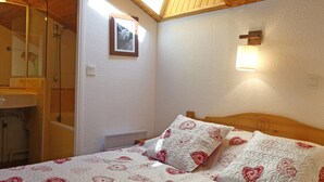 A bedroom with a wooden ceiling, a bed with patterned bedding, a bedside lamp, and a framed picture on the wall. - Charming 2-Story Duplex, Ski-In, Perfect for Families (Méribel)