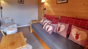 A cozy room with a wooden wall, a grey sofa with red and white checkered pillows, a small wooden table, and a lamp. - Charming 2-Story Duplex, Ski-In, Perfect for Families (Méribel)
