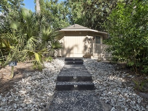 Simple, single-story building with a flat roof, surrounded by lush vegetation and a gravel path.