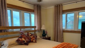 Une chambre à coucher avec une tête de lit en bois, un lit agrémenté d’oreillers à motifs, une table de chevet avec une lampe, et une vue sur l’extérieur par une fenêtre.