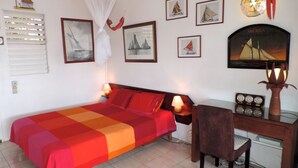 A bedroom with a bed, a desk, and a chair. There are framed pictures on the wall, a clock, and a decorative plant.