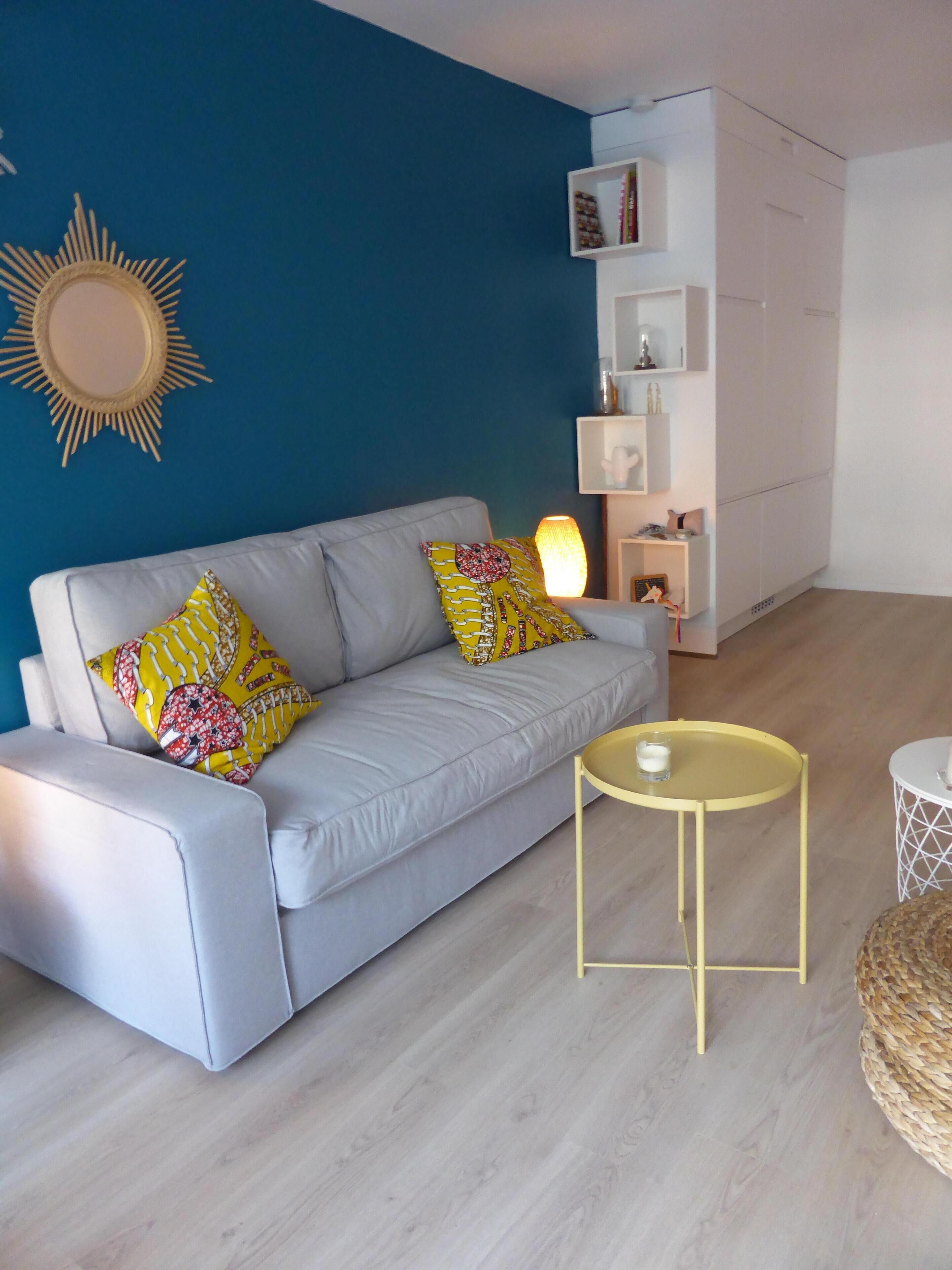 Een moderne woonkamer met een grijze bank, gele kussens met een patroon, een klein rond geel tafeltje en een witte kast met planken.