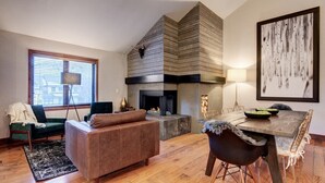 A modern living room with a fireplace, a dining table with chairs, a sofa, and a large window with blinds.