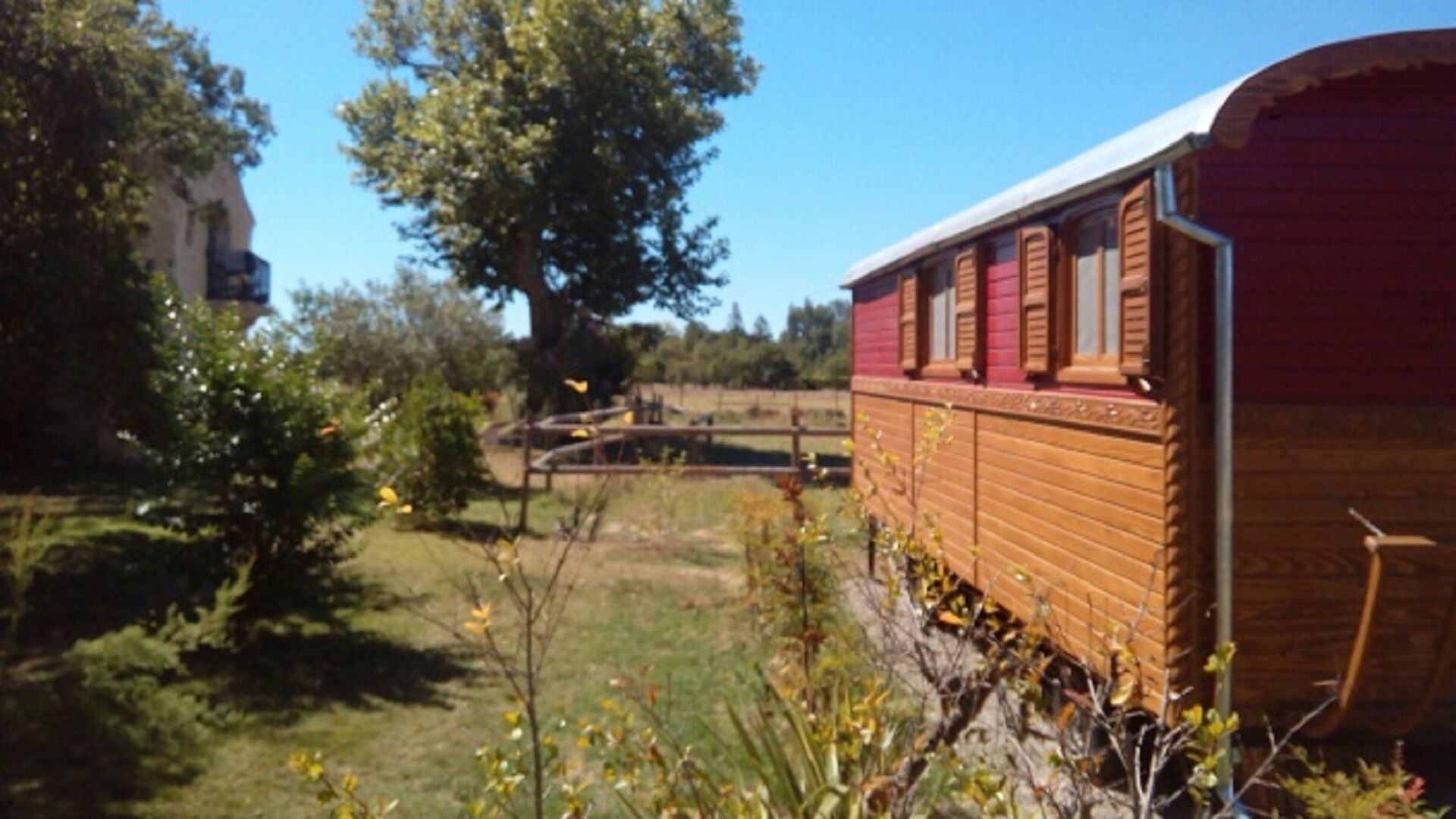 A red train car with a wooden exterior, situated next to a grassy area with trees and a fence.