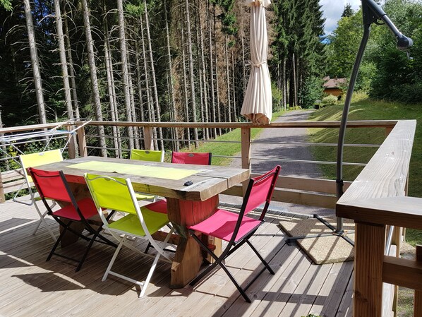 A wooden deck with a table and chairs, surrounded by a railing and overlooking a forested area.