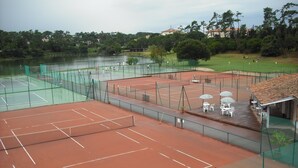 Ein Tennisplatz mit Netz, umzäumt von einem Zaun, und ein Sitzbereich mit Stühlen und Sonnenschirmen.